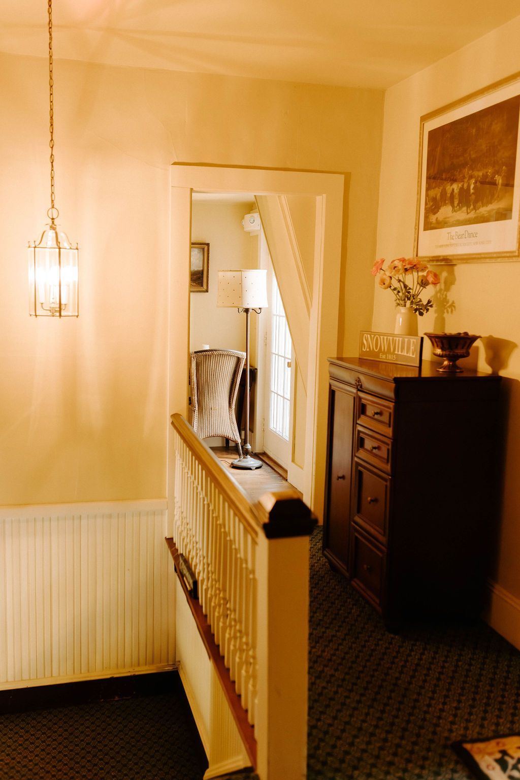 Dim hallway with cream walls, a hanging lamp, white radiator, mirror, and dark dresser with flowers.