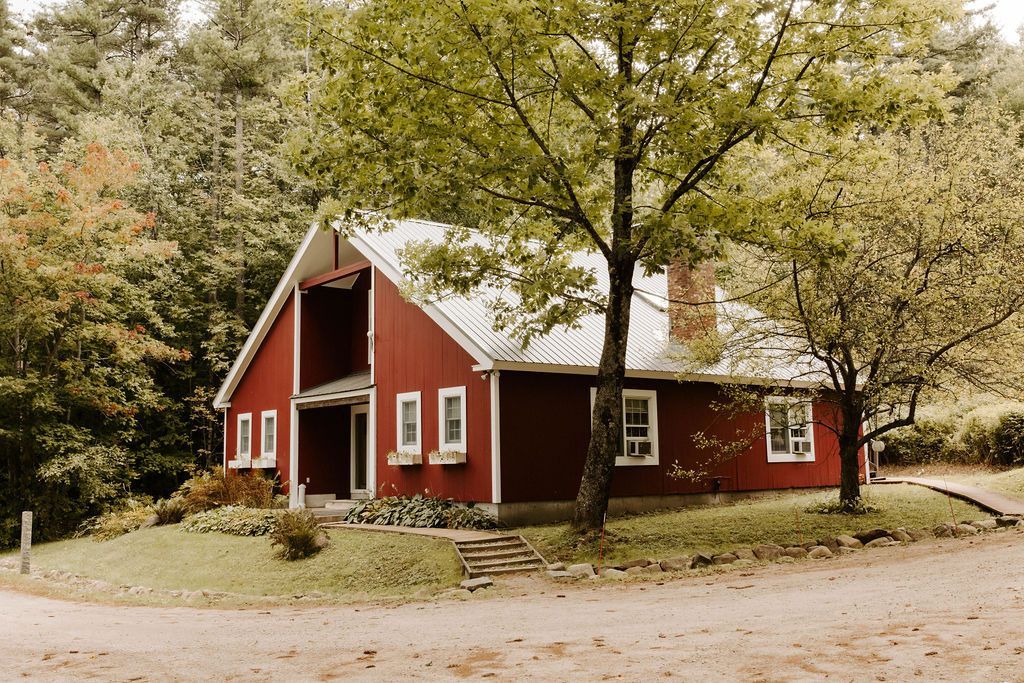 Red house with white trim under a large tree on a grassy lot, beside a curved driveway