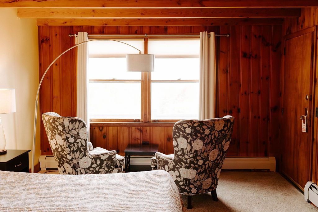 Wood-paneled cabin bedroom with floral armchairs, bed, lamp, and large window