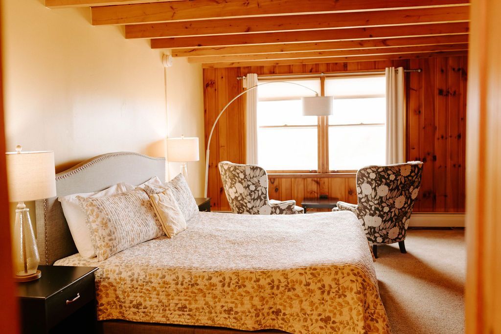 Warm bedroom with a bed, floral chairs, exposed wooden beams, and a window letting in soft light.