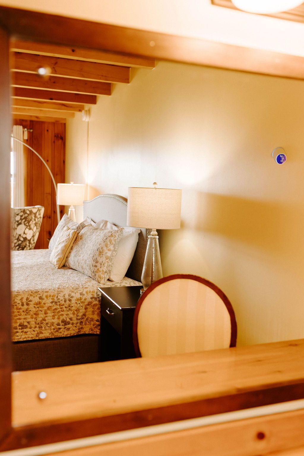 Cozy bedroom with a bed, bedside lamp, and wooden ceiling beams, viewed through a mirror.