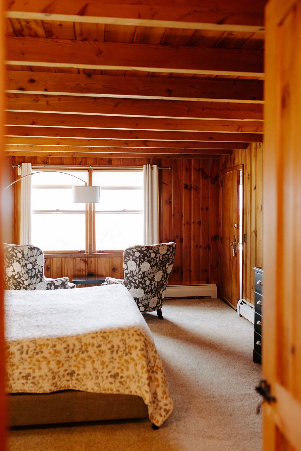 Wood-paneled bedroom with a bed, patterned chairs, and a window letting in soft light