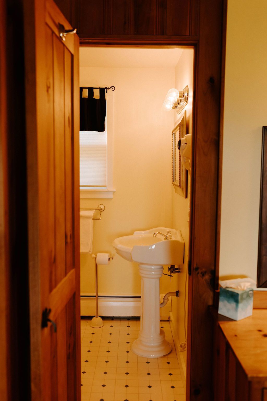 Open doorway to a small bathroom with a white pedestal sink, tiled floor, and window with dark curtain.