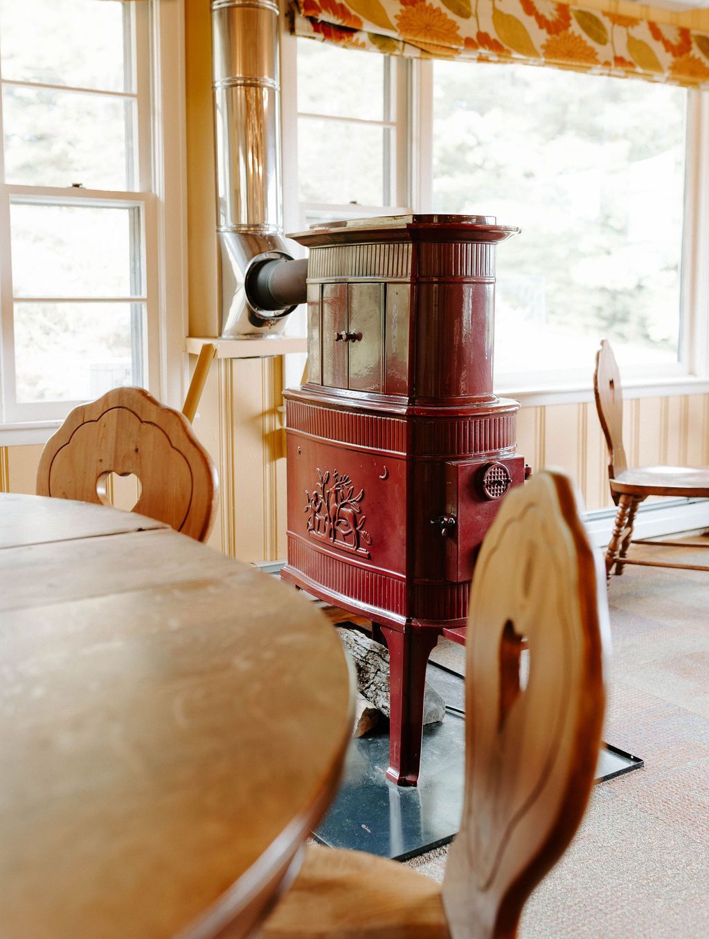 Red cast-iron wood stove in a sunlit room with wooden chairs and a table near the windows