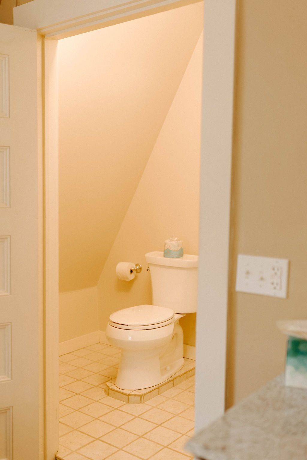 Small bathroom with a toilet, tiled floor, and beige walls, seen through an open doorway.