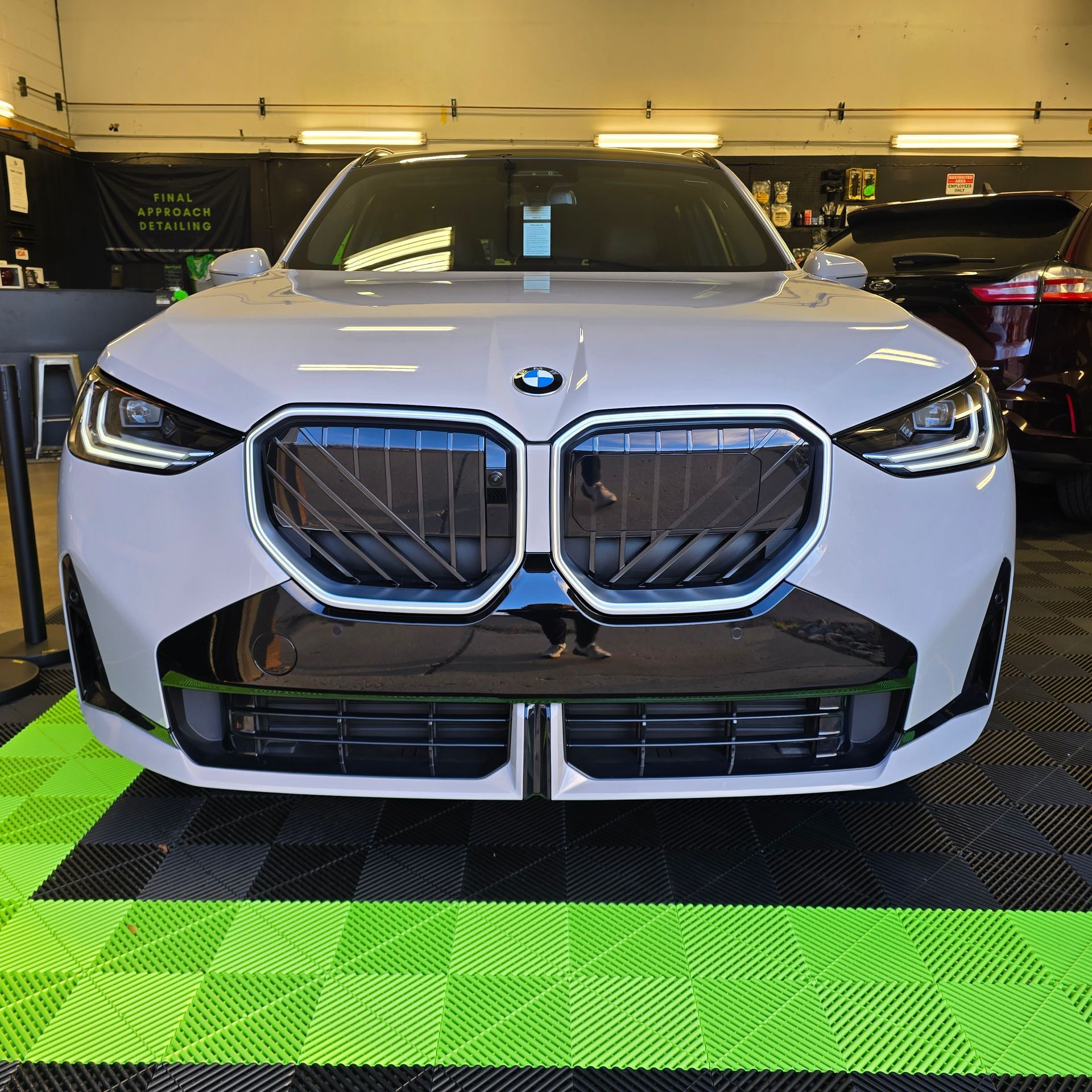 White BMW SUV, front view. Black grill, headlights, and bumper. Indoor setting with checkered floor.