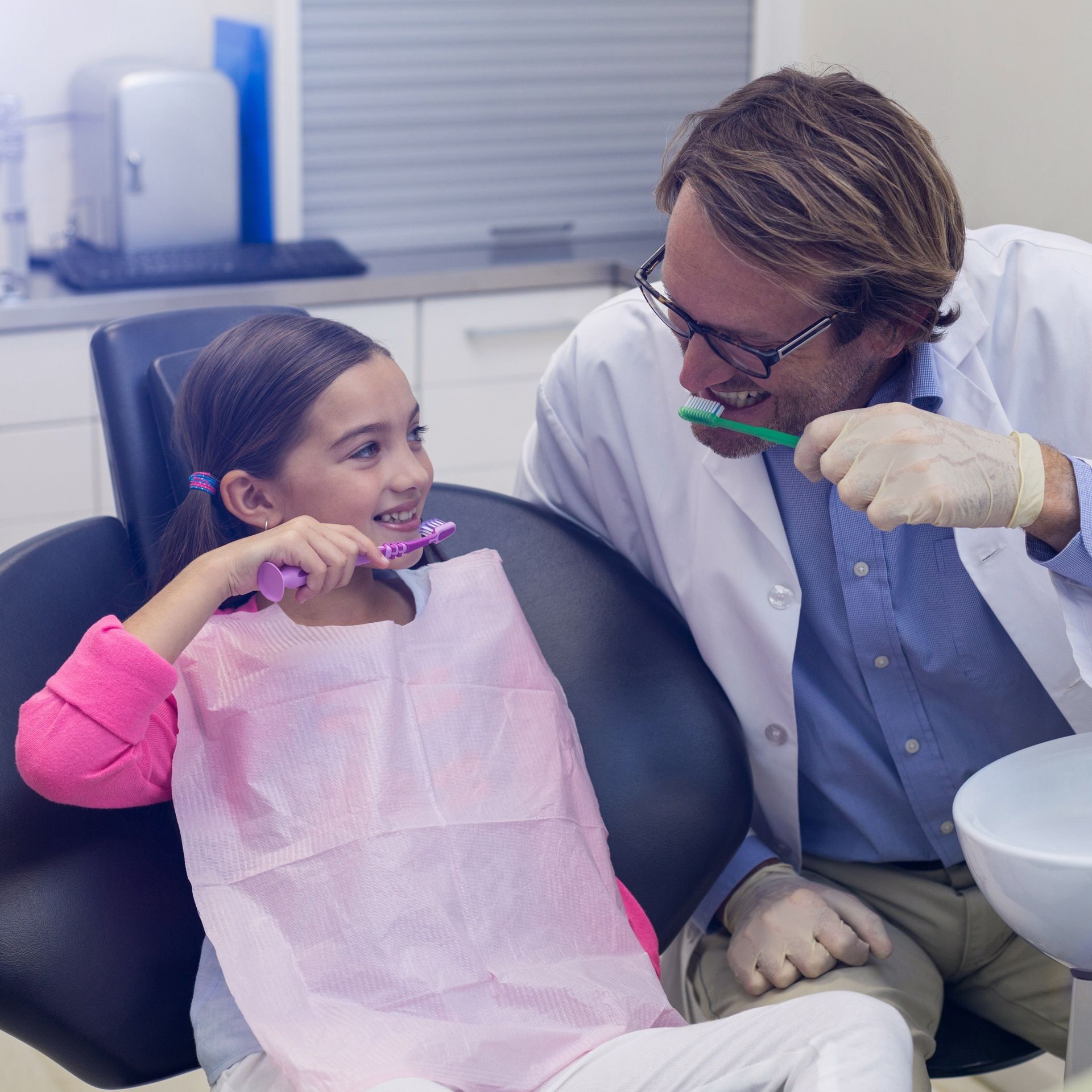 Dentist and child sitting down with toothbrushes in hand demonstrating how to brush properly