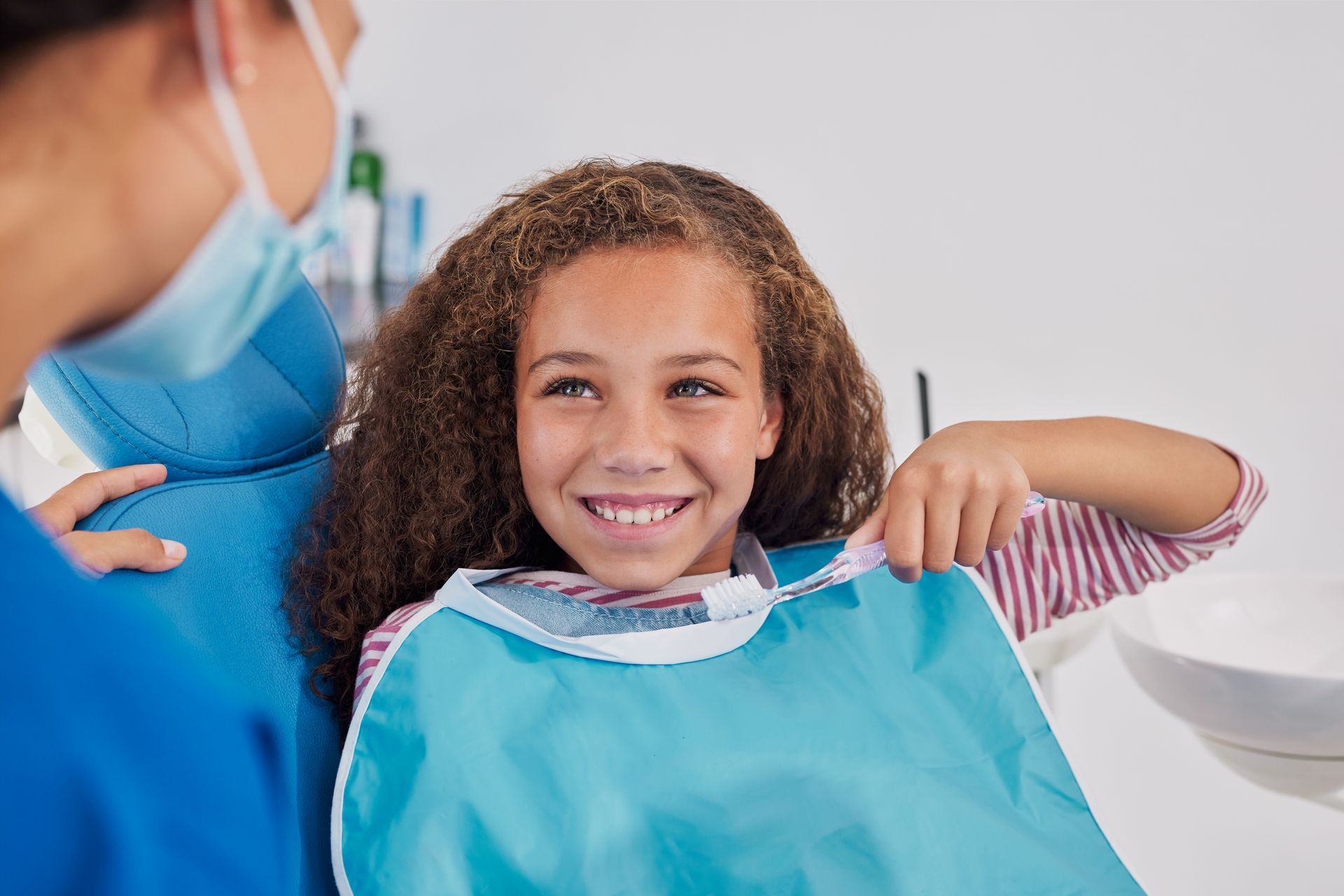 Young person smiling in dentist chair, wearing bib. Dentist in blue scrubs and mask looks on.