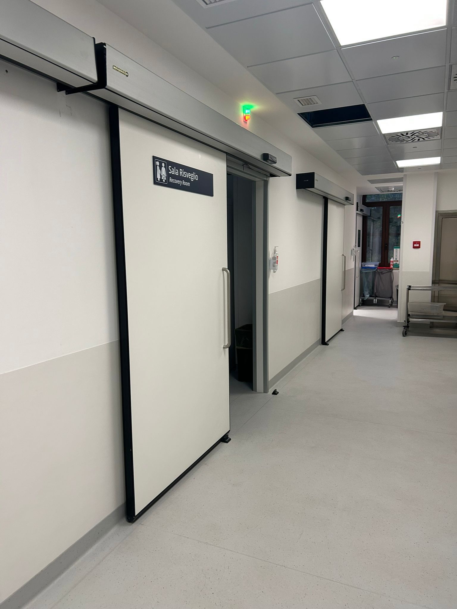 Corridoio dell'ospedale con porte scorrevoli, pareti bianche e una luce verde sopra una porta.