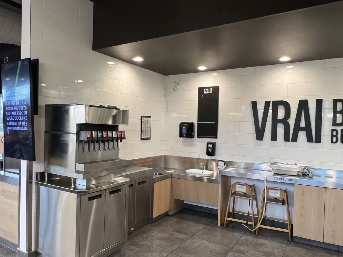 Une fontaine à soda et un comptoir à condiments dans un restaurant aux murs carrelés de blanc, aux armoires en bois et doté de deux chaises hautes.
