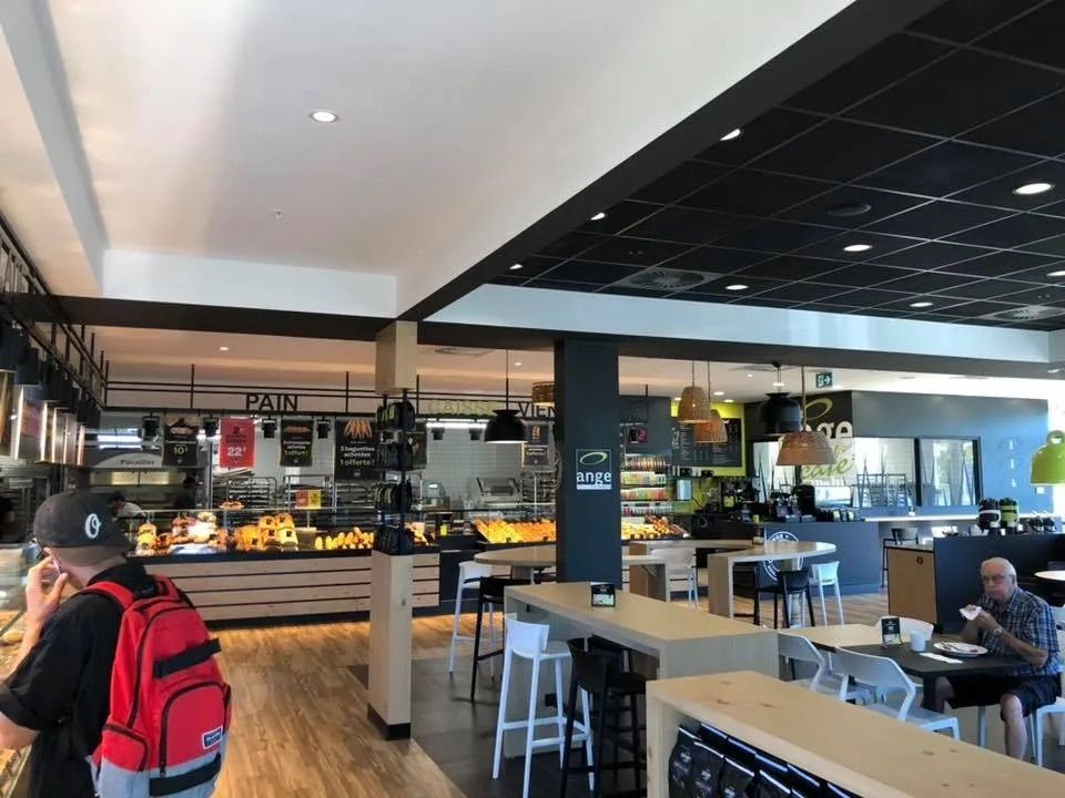 L'intérieur moderne et spacieux d'une boulangerie-café comprend des comptoirs d'exposition, des tables en bois et des tabourets sous un plafond noir.