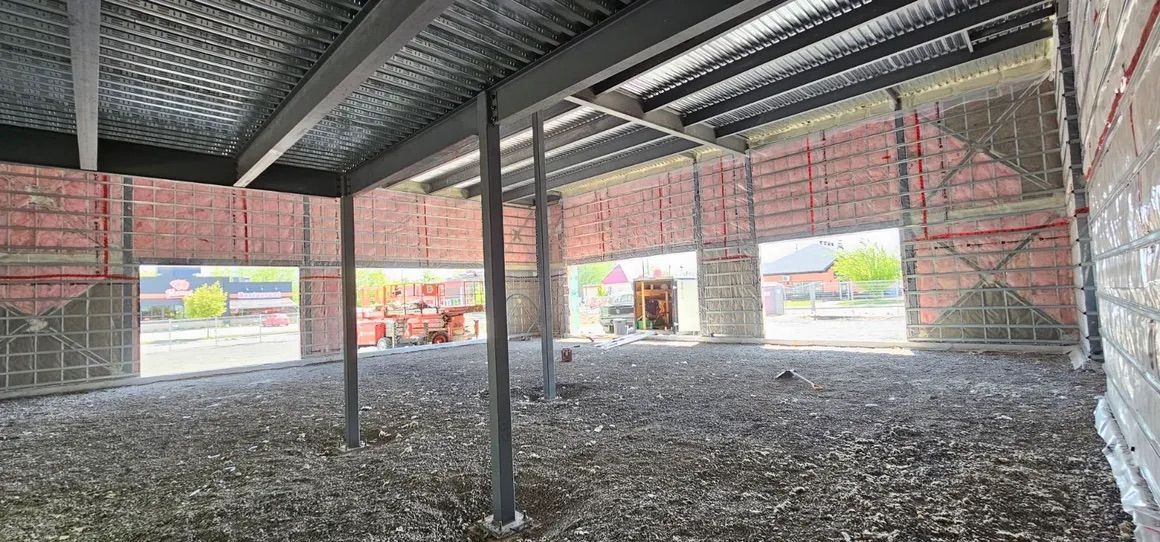 Vue intérieure d'un bâtiment en construction, montrant des poutres d'acier apparentes, des murs en briques et un sol en gravier.