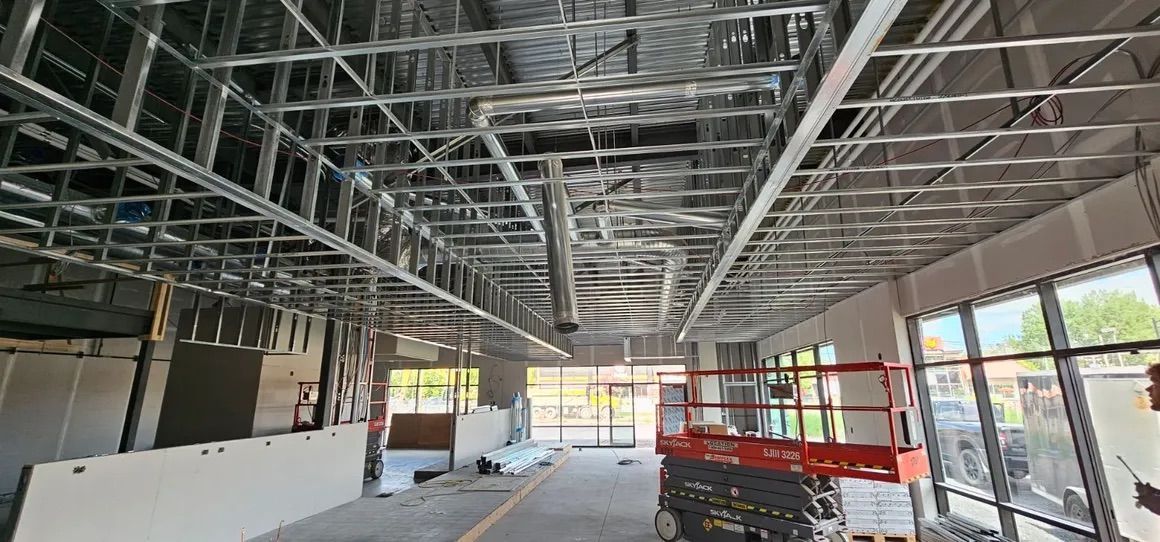 Vue intérieure d'un bâtiment commercial en construction, avec une charpente métallique apparente au plafond et une nacelle élévatrice rouge.