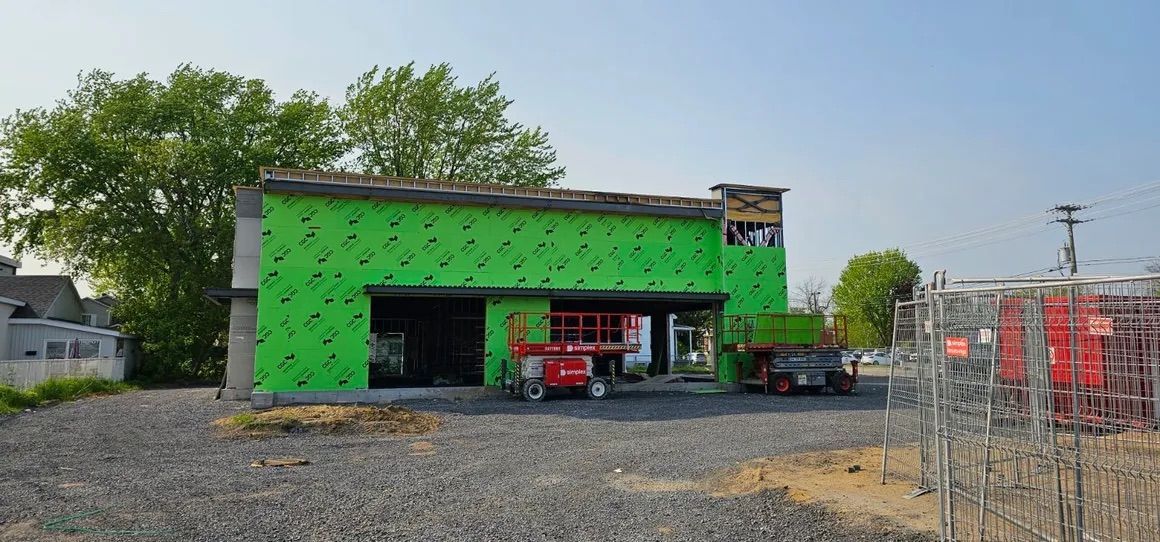 Un chantier de construction avec un bâtiment aux panneaux extérieurs vert vif, une nacelle élévatrice rouge et un sol en gravier.