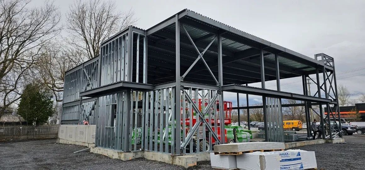 Un bâtiment commercial de deux étages en construction, avec une structure en acier apparente, des murs à ossature métallique et un sol en gravier.