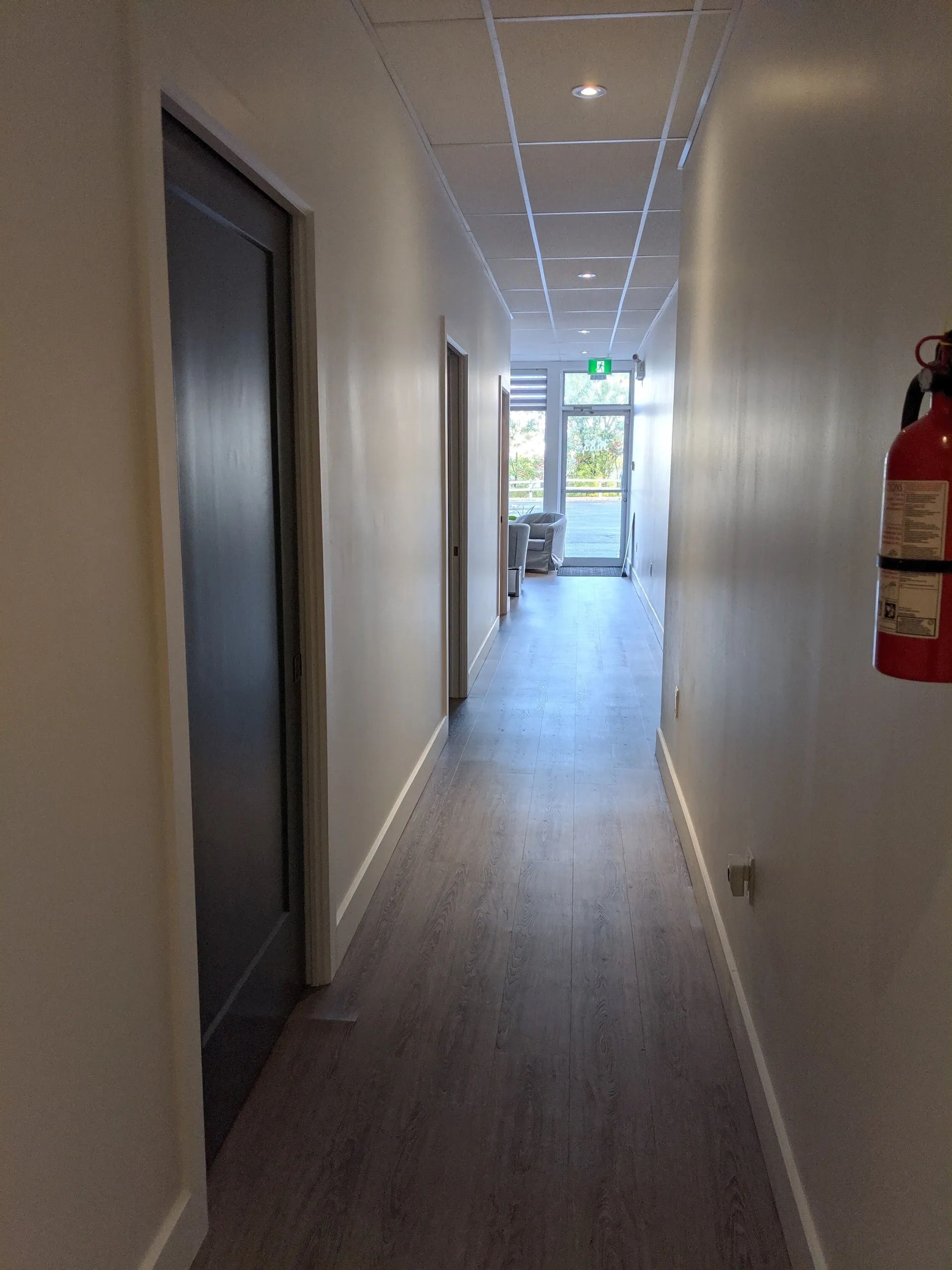 Un couloir avec un parquet clair, des murs blancs, une porte gris foncé sur la gauche et un extincteur sur la droite.
