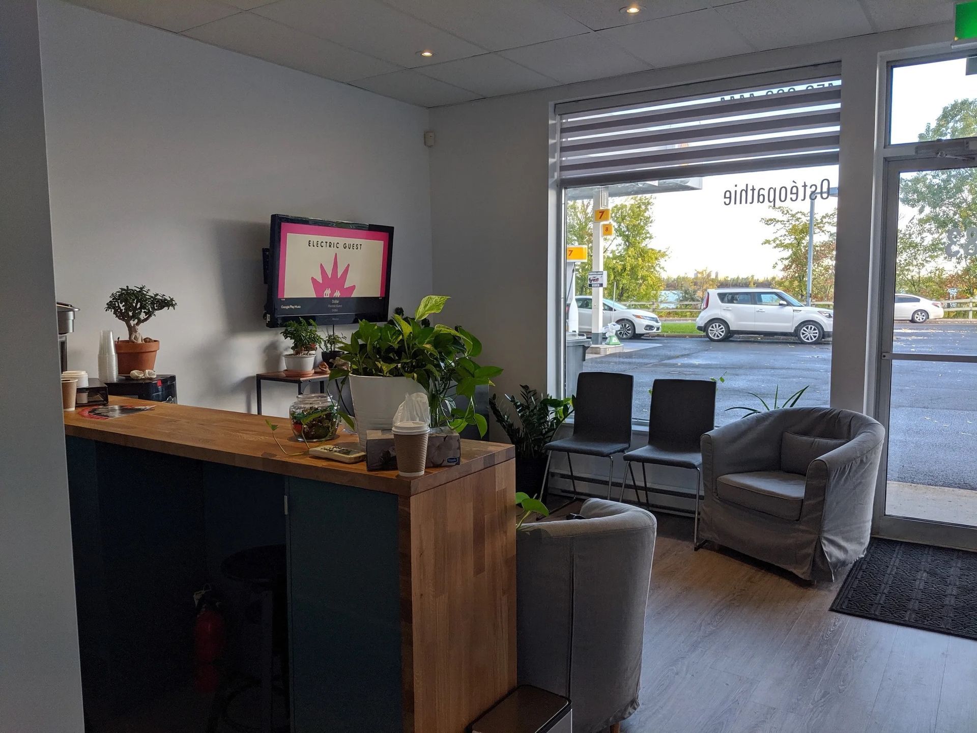 Une salle d'attente avec un comptoir d'accueil, des plantes en pot, une télévision, deux fauteuils gris et une grande fenêtre donnant sur la rue.