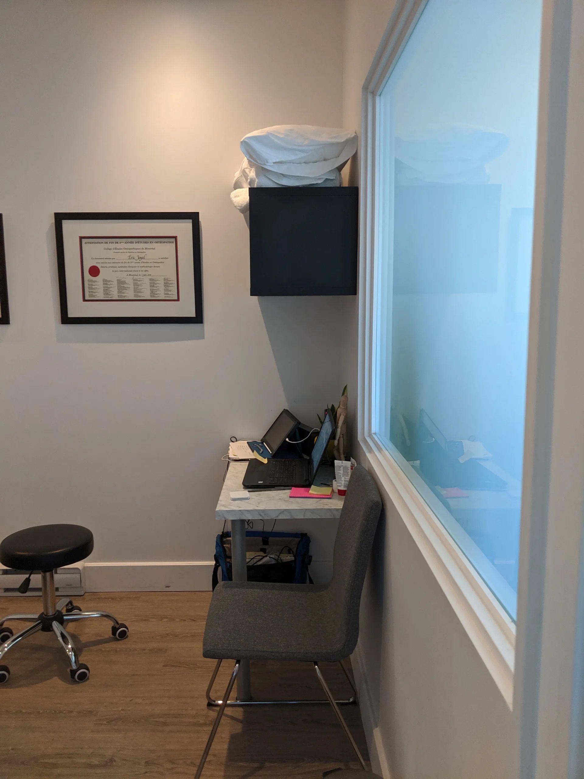 Petit espace de bureau avec un bureau, un ordinateur portable, une chaise, un tabouret à roulettes et un certificat encadré au mur.