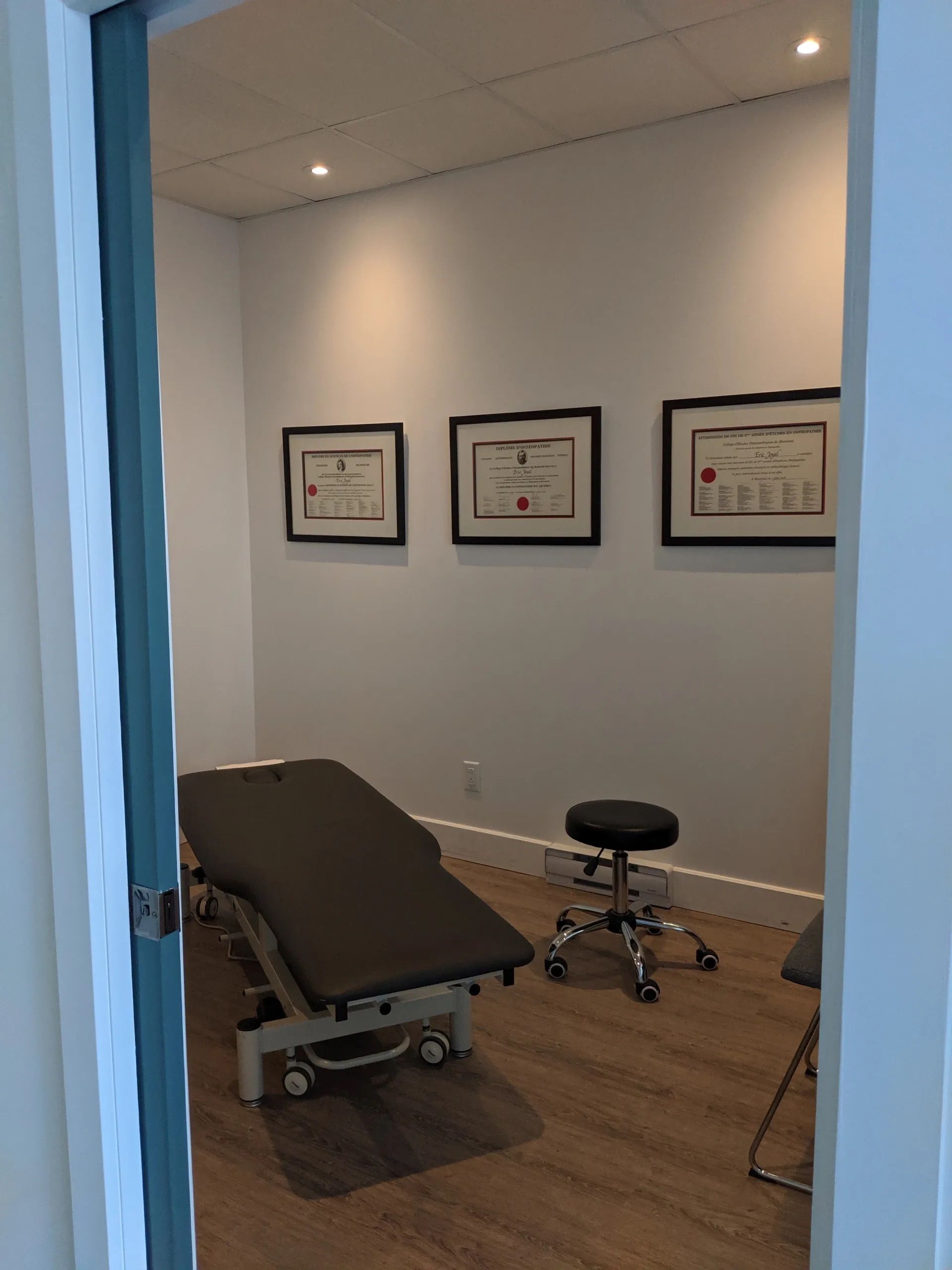 Une petite salle de soins aux couleurs neutres, équipée d'une table d'examen médical, d'un tabouret à roulettes et de trois cadres muraux.