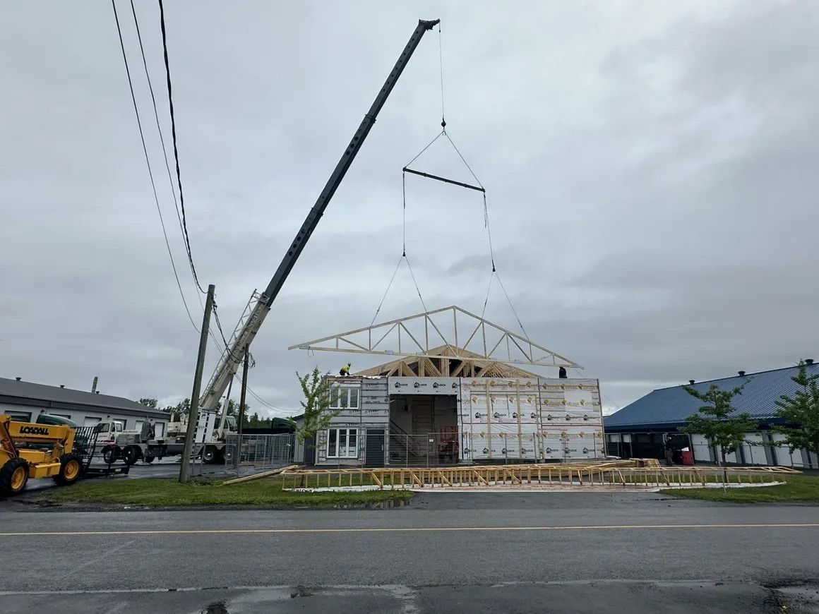 Un chantier de construction montrant une grue soulevant une grande ferme de toit en bois pour la poser sur la charpente d'un bâtiment, sous un ciel nuageux.
