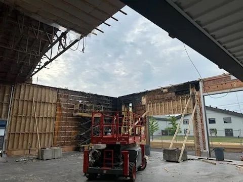 Une nacelle élévatrice rouge se dresse à l'intérieur d'un bâtiment en cours de démolition, aux murs de briques apparentes, à la charpente en bois et aux plafonds ouverts.