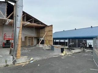 Des ouvriers construisent une nouvelle extension de bâtiment adjacente à une structure existante avec un toit en métal bleu sur un terrain pavé.