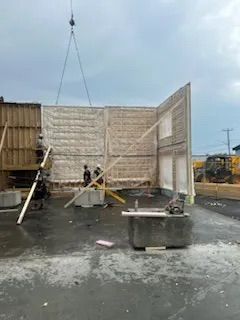 Sur un chantier, des ouvriers assemblent de grands panneaux muraux en béton, mis en place par une grue.