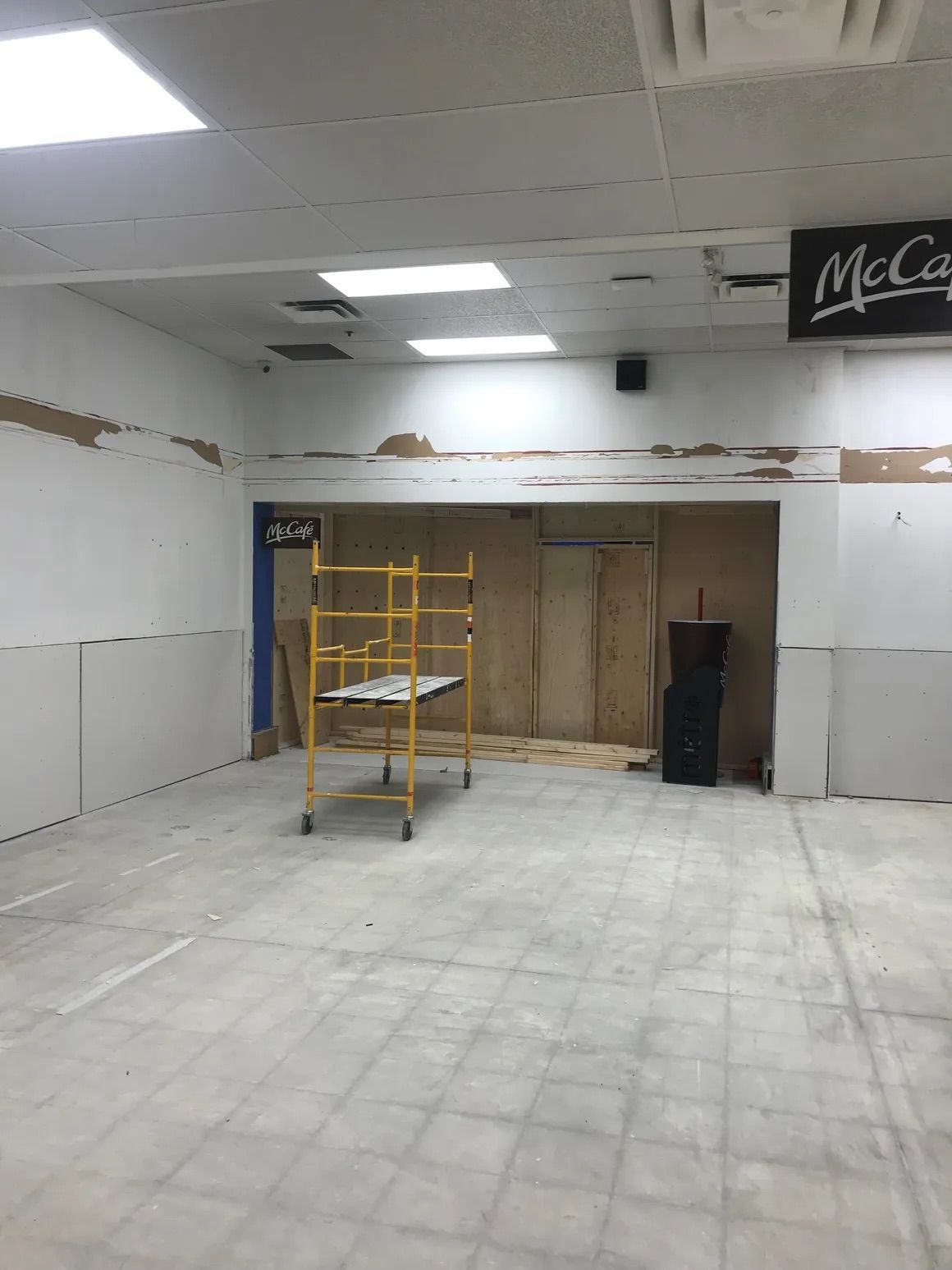 Vue intérieure d'une pièce en construction, avec des murs en plaques de plâtre blanches, un sol en béton et un échafaudage jaune.