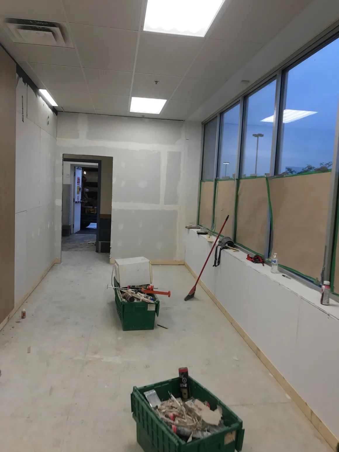Une pièce intérieure en travaux, avec des cloisons sèches apparentes, de grandes fenêtres recouvertes de papier kraft et des caisses vertes au sol.