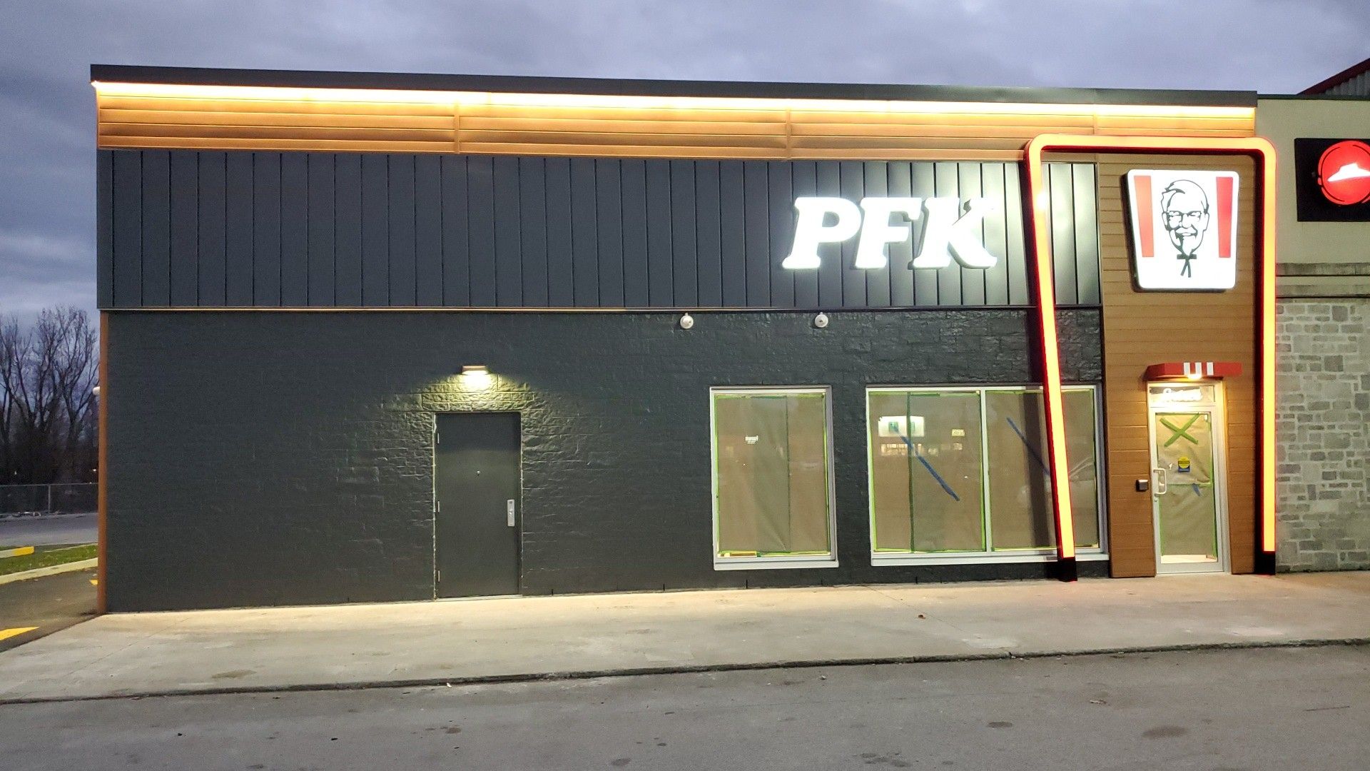 La façade moderne d'un restaurant PFK, avec ses murs gris foncé, son entrée lambrissée et son enseigne illuminée au crépuscule.