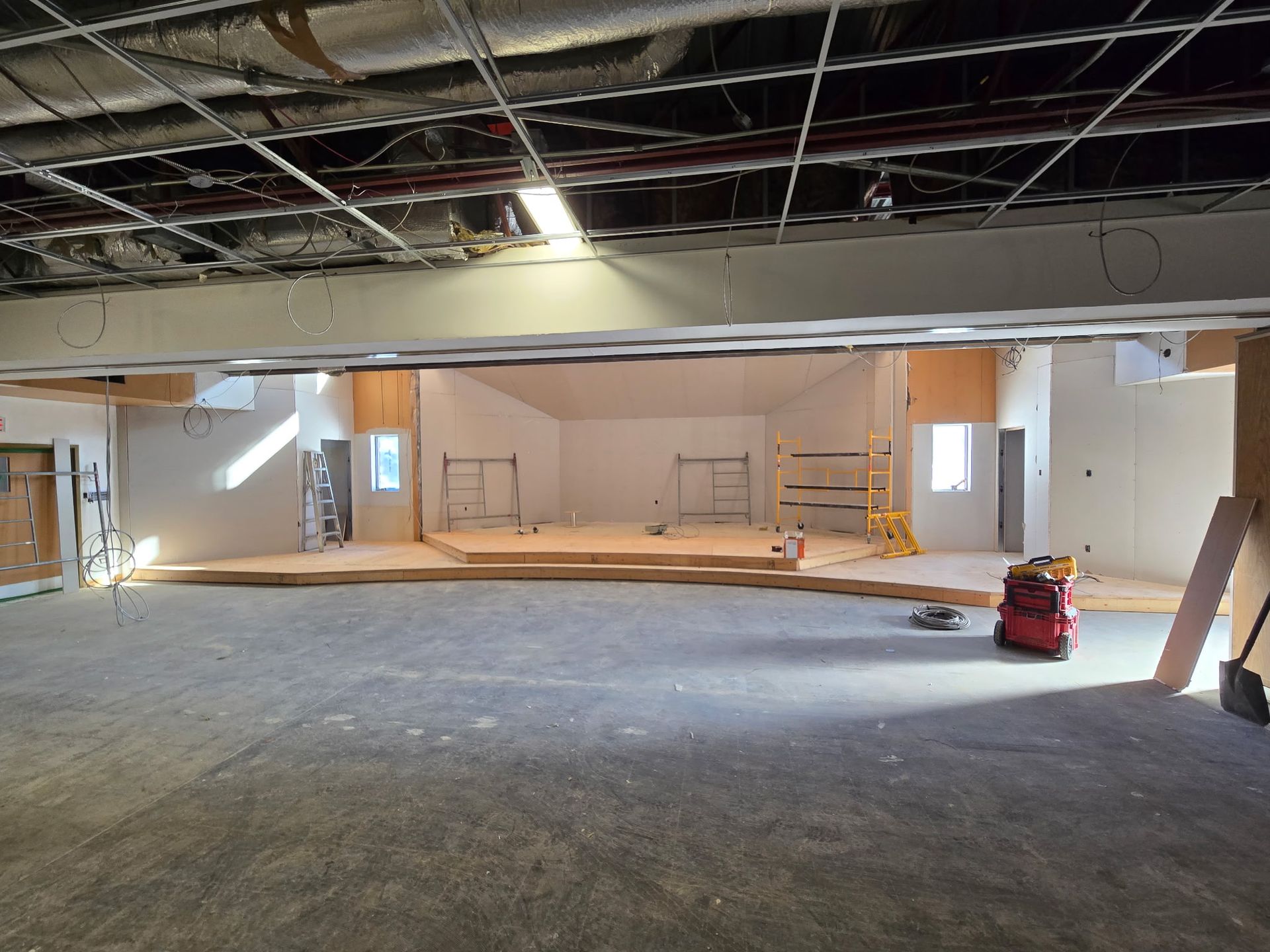 Intérieur d'un bâtiment en rénovation, comprenant une scène surélevée, des murs inachevés et des tuyaux apparents au plafond.