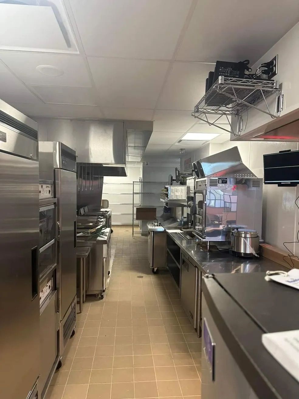 Une cuisine commerciale étroite avec des appareils électroménagers en acier inoxydable, des plans de travail et un sol carrelé.