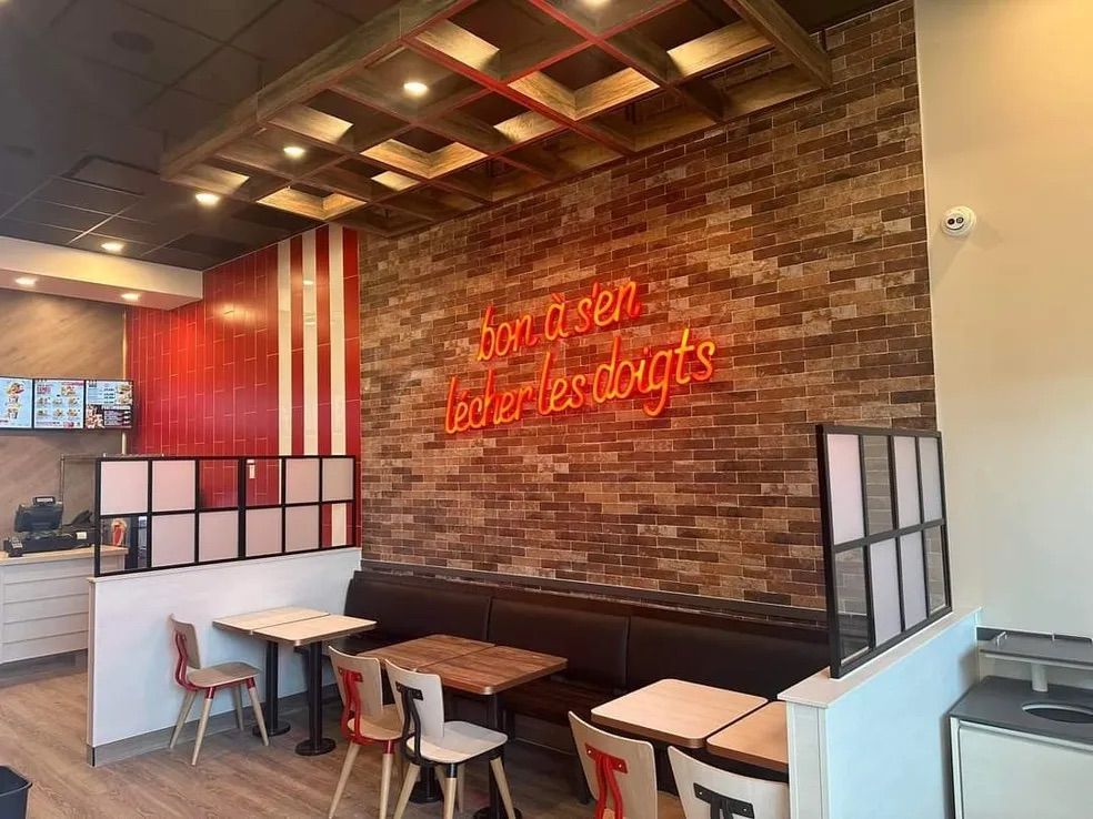A KFC interior with wooden tables, dark booths, and a neon sign on a brick wall reading, 