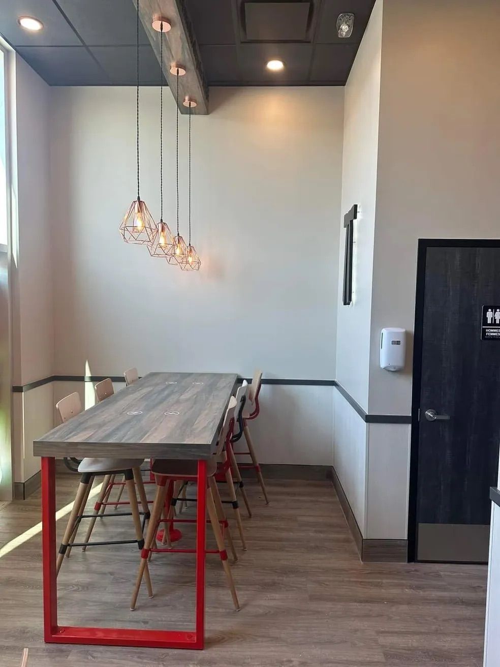 Dans une pièce aux murs clairs, au parquet et aux suspensions modernes, une table haute en bois avec un cadre en métal rouge est placée.