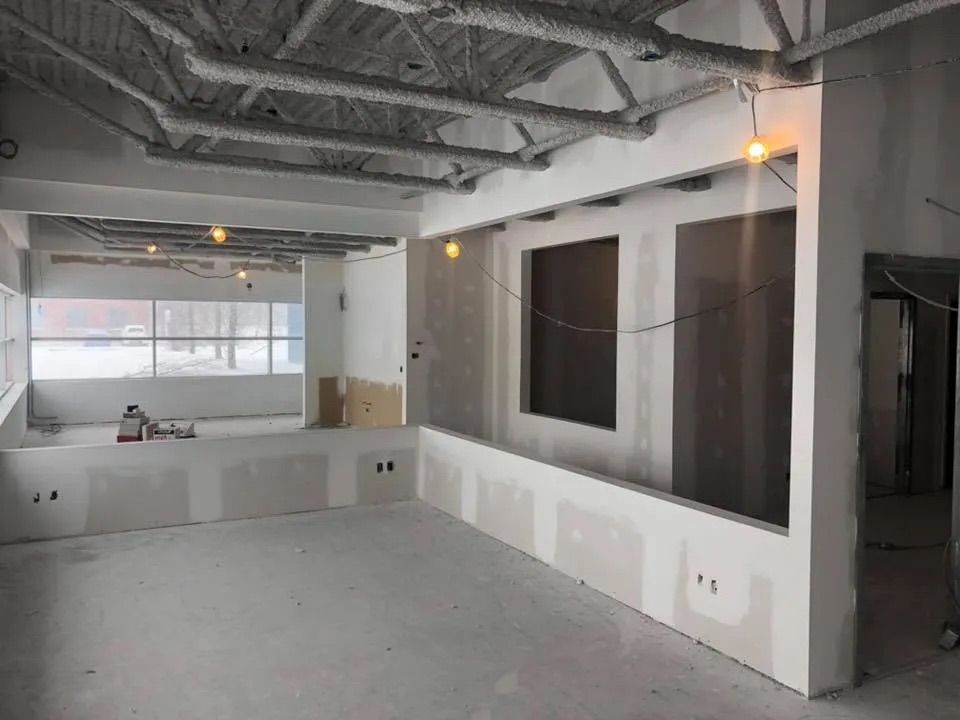 Scène de construction intérieure d'une pièce avec des cloisons sèches non finies, des poutres apparentes au plafond et de grandes fenêtres.