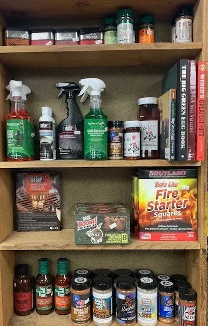 A wooden shelf filled with grilling supplies: spice jars, sauces, sprays, fire starter, and cookbooks.
