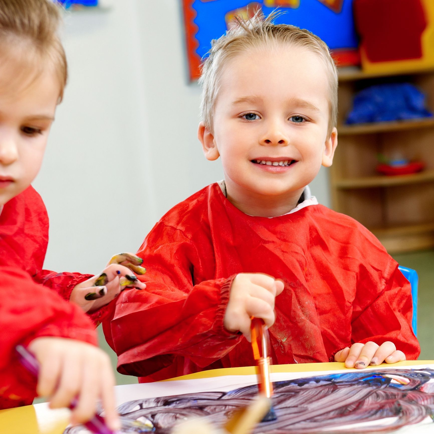 Smiling young boy painting at a table, wearing a red smock, inside a classroom setting. Smiling young boy painting at a table, wearing a red smock, inside a classroom setting.