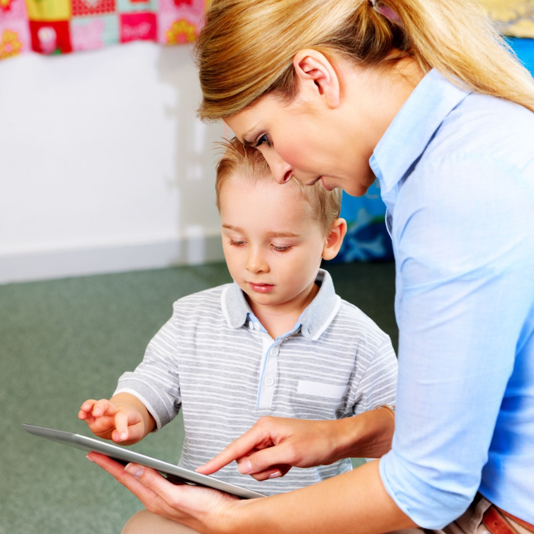 Woman in blue shirt helps a young boy in a grey shirt use a tablet indoors. Woman in blue shirt helps a young boy in a grey shirt use a tablet indoors.