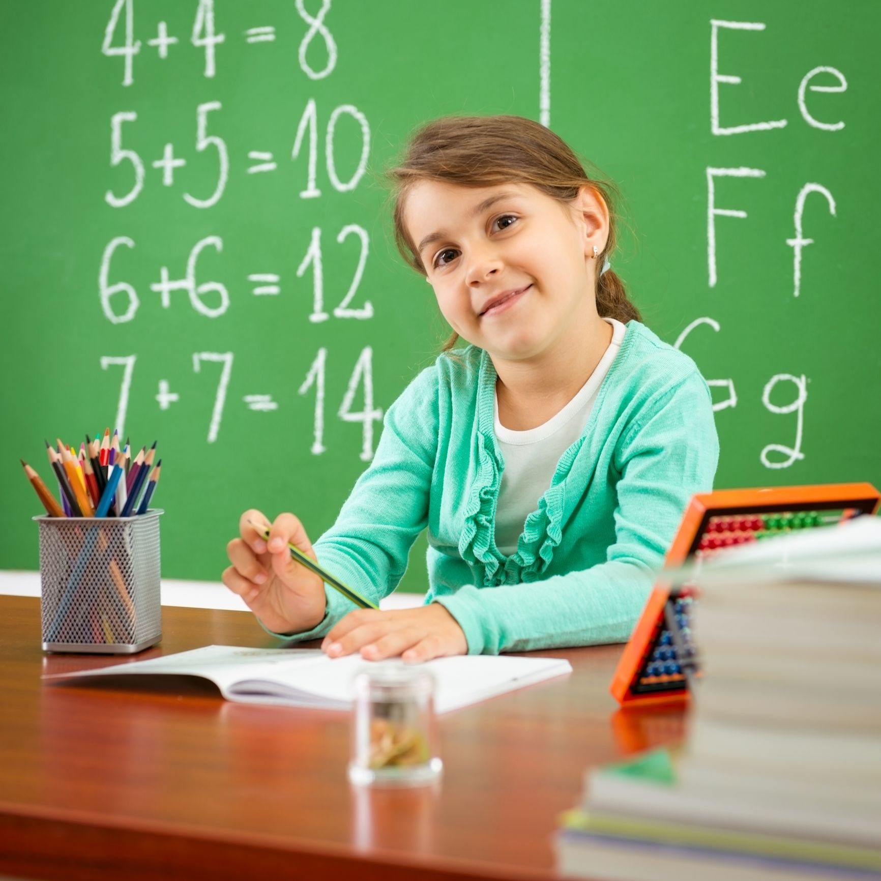 Girl in teal cardigan smiles, writing at desk in front of chalkboard with math problems. Girl in teal cardigan smiles, writing at desk in front of chalkboard with math problems.
