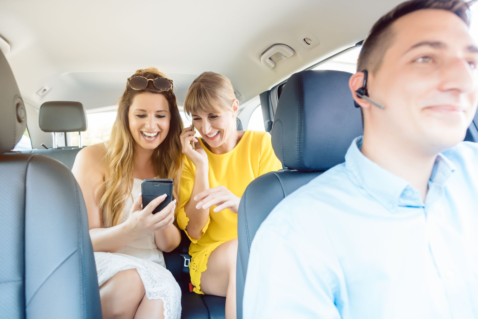 Two women in a car looking at a phone, driver in the front wearing a headset.