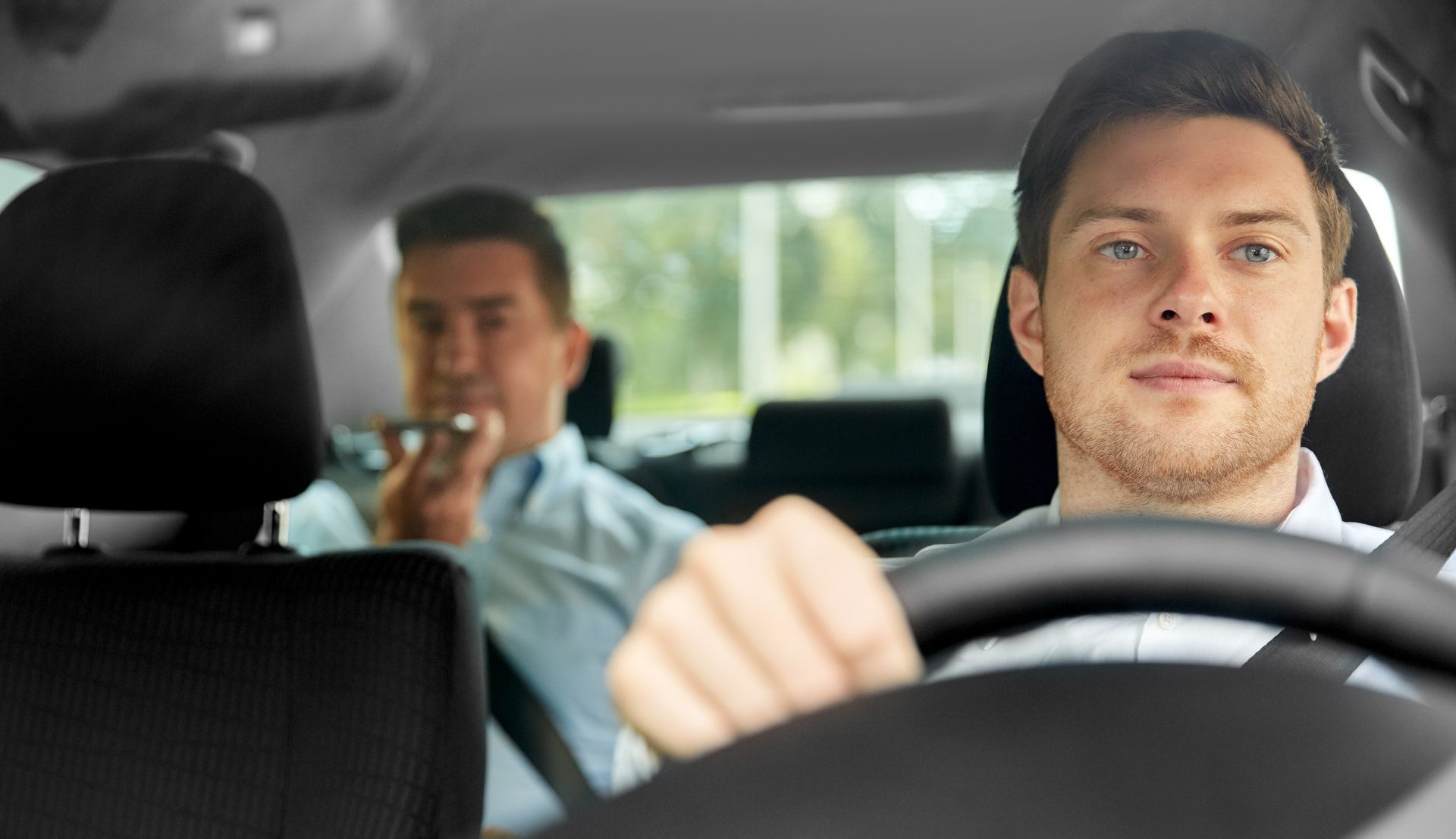 Man driving car, passenger in back seat, light background.