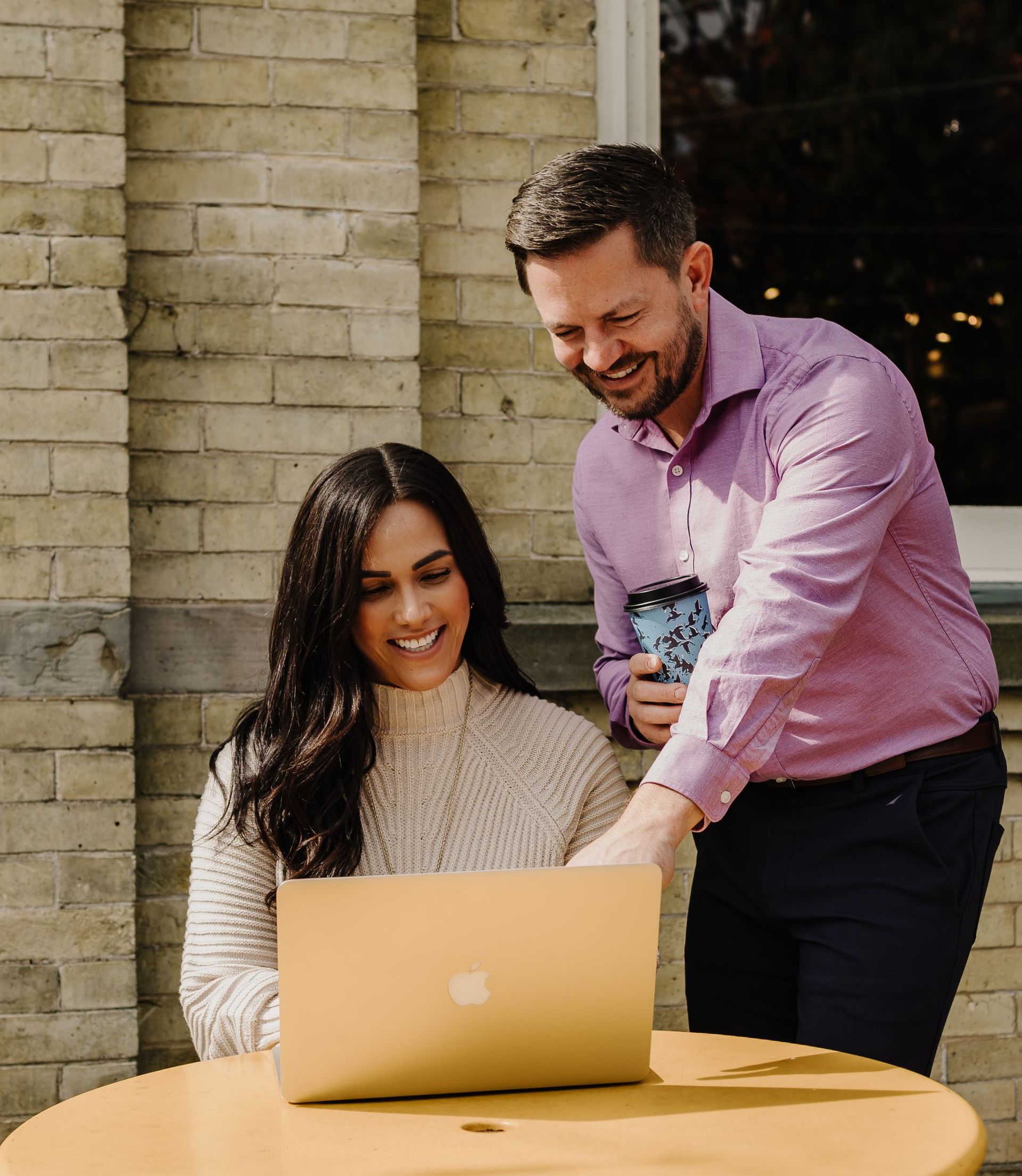 Woman and man looking at a laptop together, smiling. Man points at the screen. Outdoors by a brick wall.