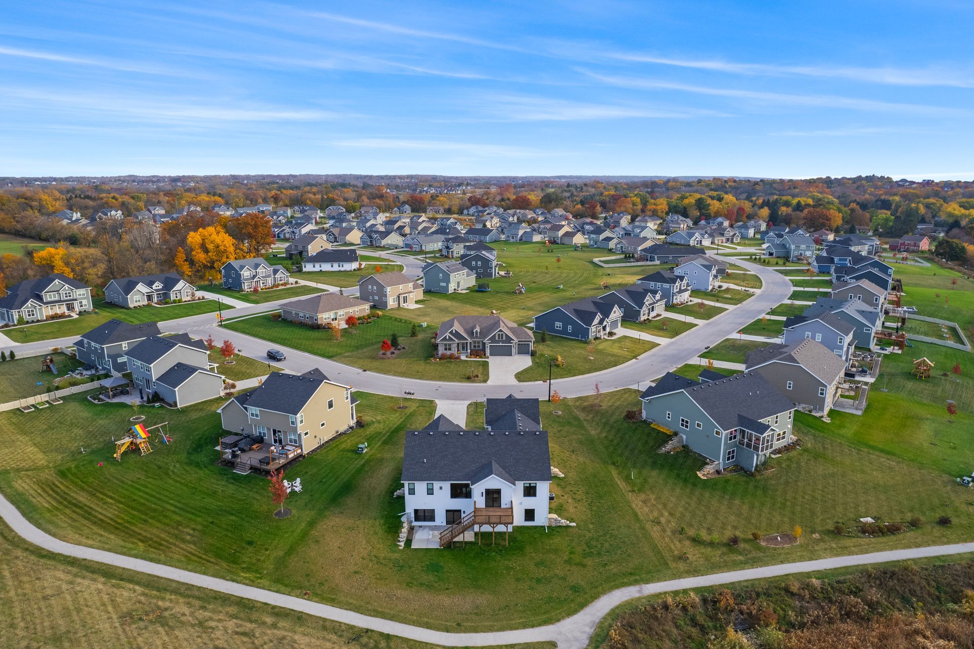 Aerial view of a suburban neighborhood with houses surrounding a curving road, green lawns, and a blue sky.