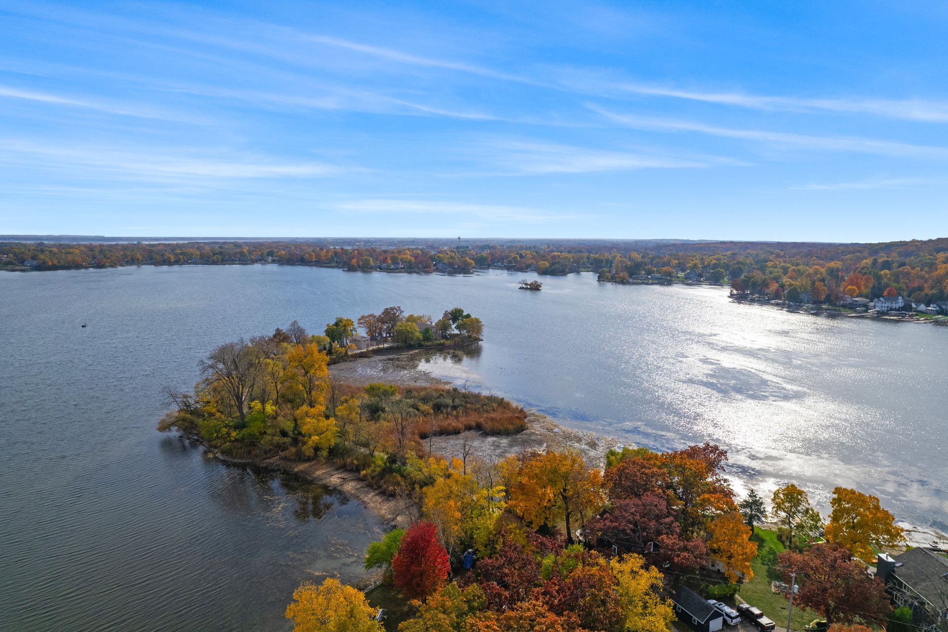 Aerial view of a lake with an island in the foreground surrounded by trees with fall colors under a blue sky.