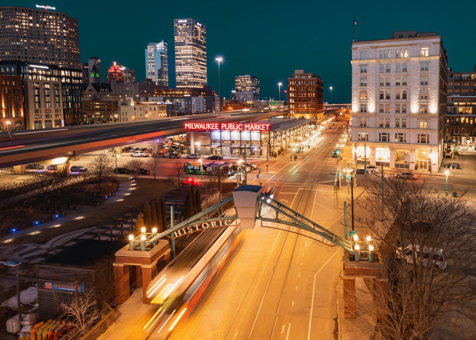 Third Ward Milwaukee city street at night with lit buildings, bridge, and a passing train.