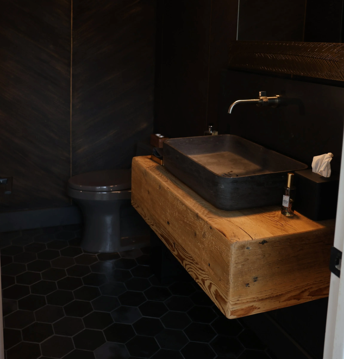 Dark modern bathroom with a wooden sink vanity, black countertop, and toilet on hexagonal tile floor