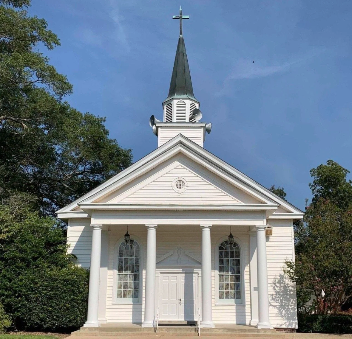 Small white church with a tall steeple and cross, framed by trees under a blue sky