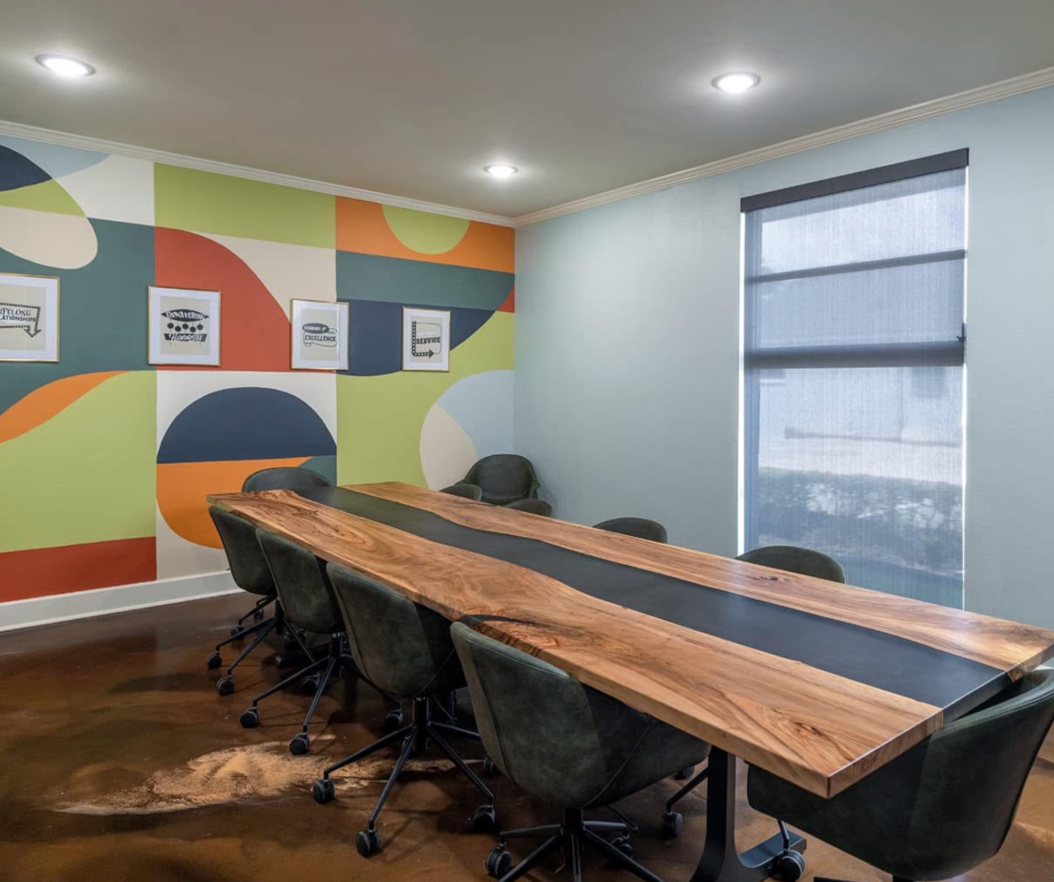 Modern conference room with a long wooden table, black chairs, colorful mural wall, and windows.
