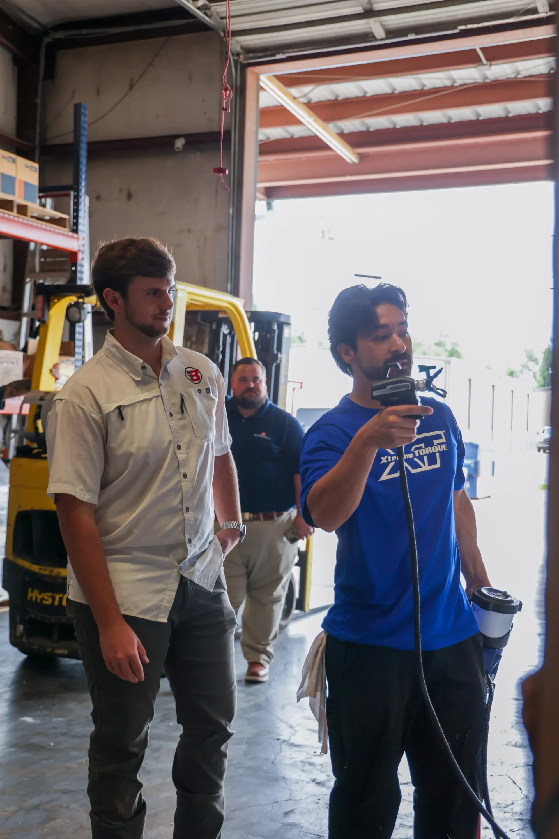 Two men standing in a workshop; one in a blue shirt holds a cable near an open garage door.