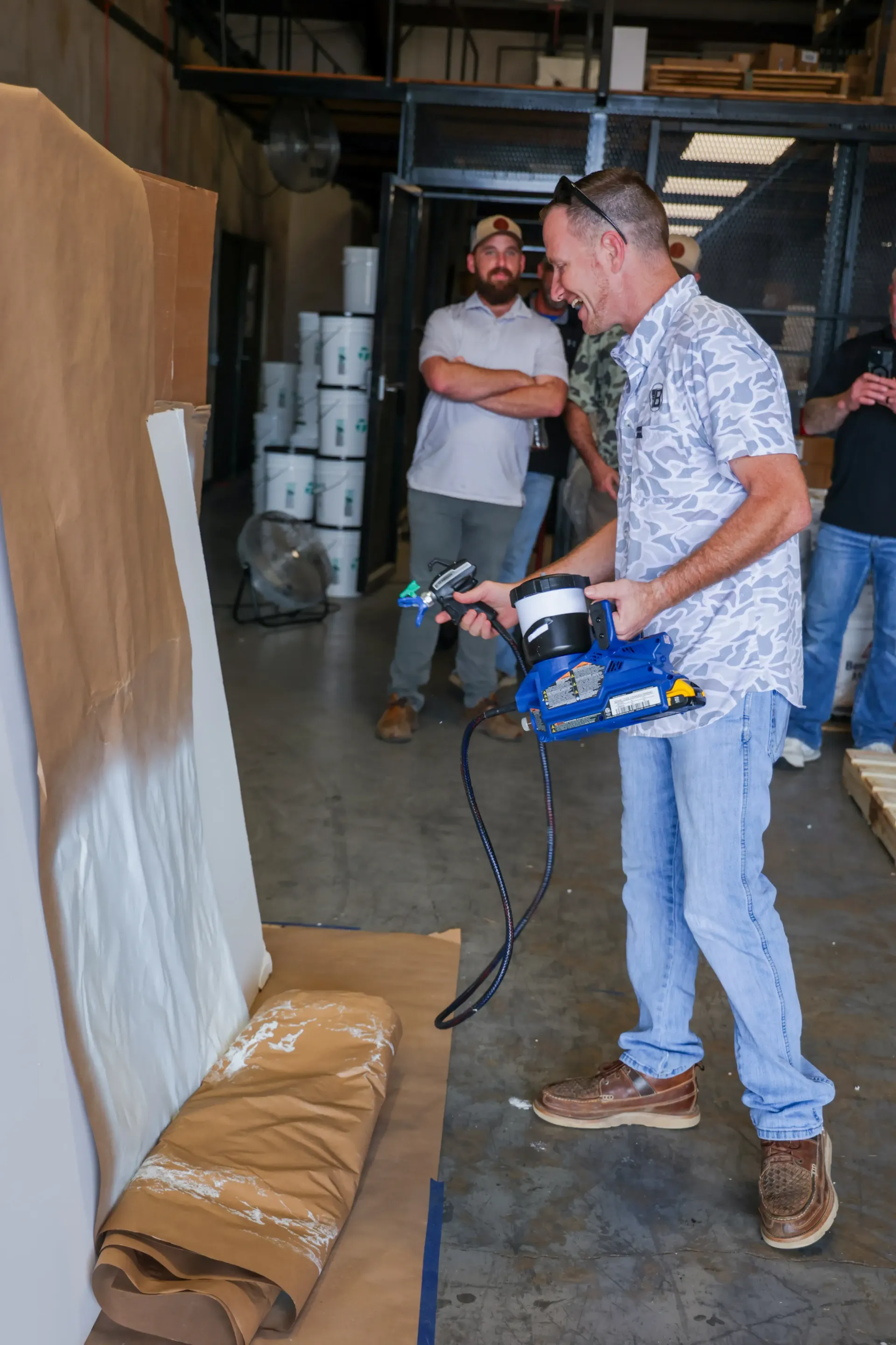 Man in a warehouse using a spray gun to coat a large wooden panel, with a second man nearby.